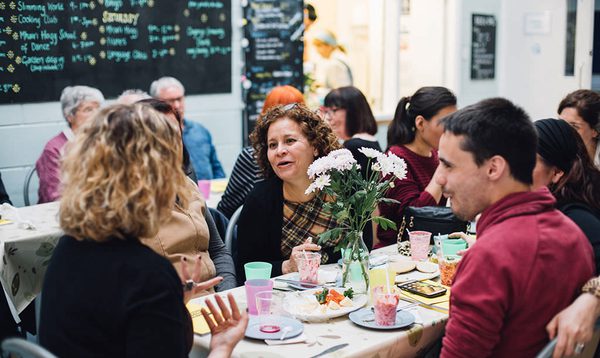 Group of people sat at a table talking with flowers in the middle and plates and cups on the table