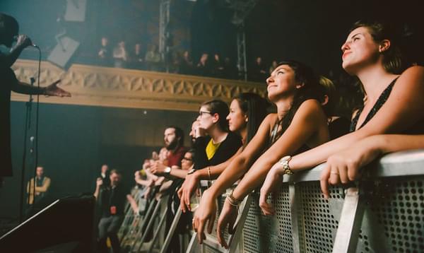 Crowd behind barrier at a concert with lights from the stage illuminating their faces