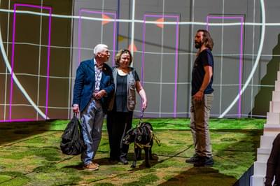 Couple on stage with a guide dog, walking around the set with background projections and grass on the floor