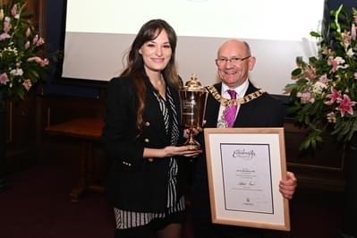 The Lord Provost Robert Aldridge, a man wearing glasses and a gold chain, is standing next to Nicola Benedetti. The Lord Provost is holding a wooden frame with The Edinburgh Award certificate inside. Nicola Benedetti is holding a Gold cup.