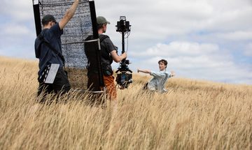 Two camera men stand in a field of corn filming a dancer in a blue shirt
