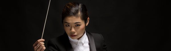 The conductor shown holding her baton and looking up at the camera, dressed in a white shirt and black tuxedo jacket.