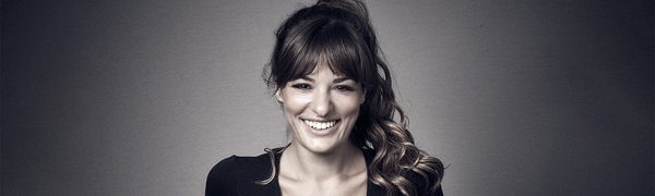 The violinist pictured standing facing the camera and smiling widely with her hands in her pockets, dressed in black against a black backdrop.