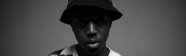 Moses Boyd poses in a black hat and black-and-white checked sweater