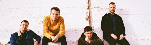 The four members of Tide Lines sit outside, leaning against a white brick wall