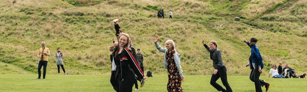 A row of smiling people perform a dance in a field, with Arthur's Seat in the background.