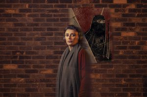 A woman wearing headphones stands facing the camera, in front of brick wall featuring a painting of a woman wearing a niqab.