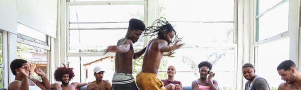 A group of young people sit around on sofas as two young men dance in the middle
