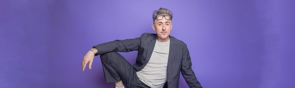 Alan Cumming pictured sitting on the floor and leaning back on one hand, with his other hand draped across his raised knee.