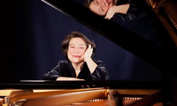 The pianist shown seated at an open grand piano, leaning forward and resting her head in her left hand. Her face is reflected in the shiny piano cover.