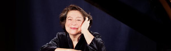The pianist shown seated at an open grand piano, leaning forward and resting her head in her left hand. Her face is reflected in the shiny piano cover.