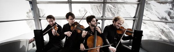 The Goldmund Quartet pictured playing their instruments seated close together in front of a window looking out on a snowy mountain scene.