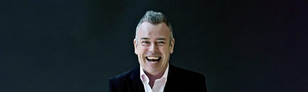 The pianist pictured wearing a white shirt and black suit jacket, standing against a black backdrop smiling widely at the camera.