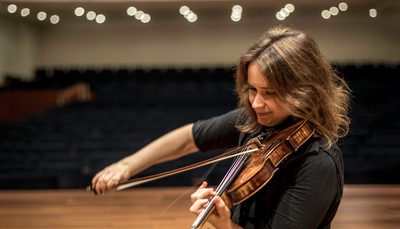 The musician shown seated cross-legged on a stage in an empty theatre, dressed in black and playing the violin.
