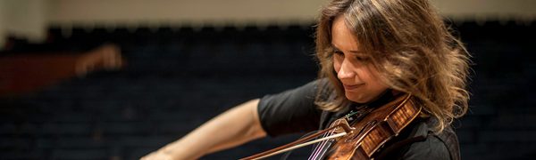 The musician shown seated cross-legged on a stage in an empty theatre, dressed in black and playing the violin.