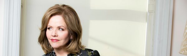 Renée Fleming, pictured leaning against a window and smiling as she gazes out, wearing a black silk shirt with a green and silver pattern.