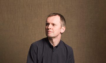 The pianist photographed sitting casually on a stool, wearing a dark grey shirt and gazing into the middle distance to the right of the camera.