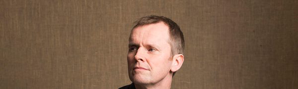 The pianist photographed sitting casually on a stool, wearing a dark grey shirt and gazing into the middle distance to the right of the camera.