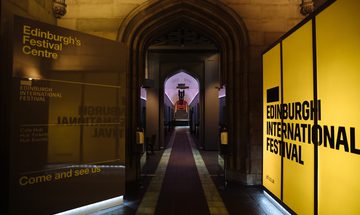 A view of a corridor with an arched roof, illuminated on one side by a bright yellow light panel reading Edinburgh International Festival and by a purple light at the far end.