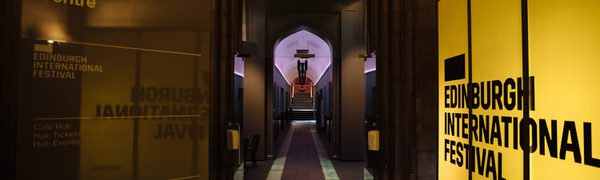 A view of a corridor with an arched roof, illuminated on one side by a bright yellow light panel reading Edinburgh International Festival and by a purple light at the far end.