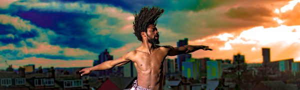 A man with dreadlocks wearing a skirt and trousers made of white fabric jumps in the air with his arms outstretched.