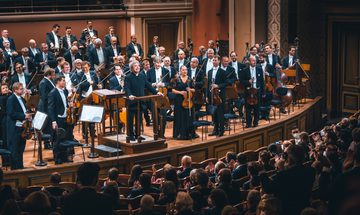 On orchestra and conductor wearing black suits and black evening dress stand on stage in front of a sitting audience.