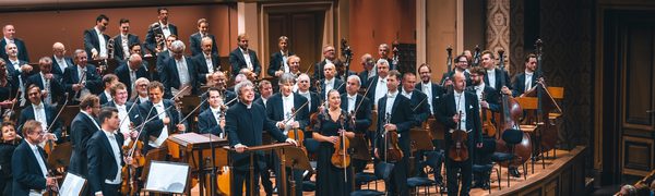 On orchestra and conductor wearing black suits and black evening dress stand on stage in front of a sitting audience.