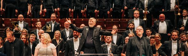An orchestra, singers and choir stand on stage with a conductor on the podium gesturing triumphantly towards the audience