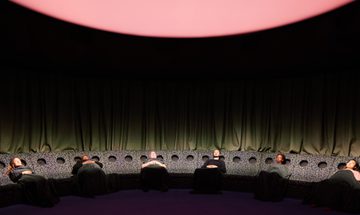 A group of people in a semicircle of reclined seating lie back with their eyes shut, with a large glowing light above them.
