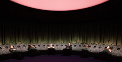 A group of people in a semicircle of reclined seating lie back with their eyes shut, with a large glowing light above them.