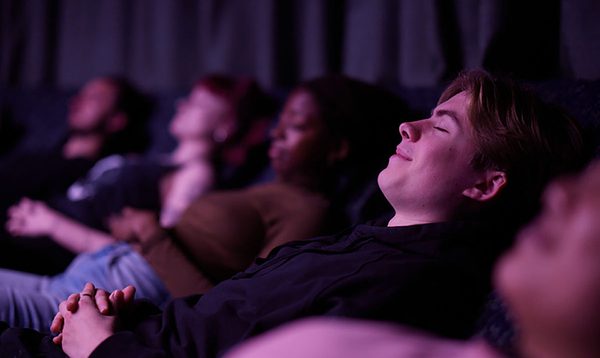 A row of reclined people lie back with their eyes closed and hands folded over their stomachs, with one woman's smiling face in focus.