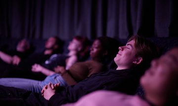A row of reclined people lie back with their eyes closed and hands folded over their stomachs, with one woman's smiling face in focus.