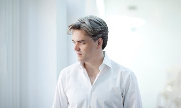The conductor pictured wearing a white shirt and looking away from the camera into the distance, photographed against a bright white background.