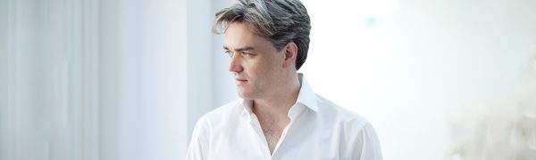 The conductor pictured wearing a white shirt and looking away from the camera into the distance, photographed against a bright white background.