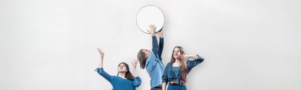 Five people dressed in blue tones are in different positions against a white wall, with a man at the back of the group holding a wide, flat drum above his head.