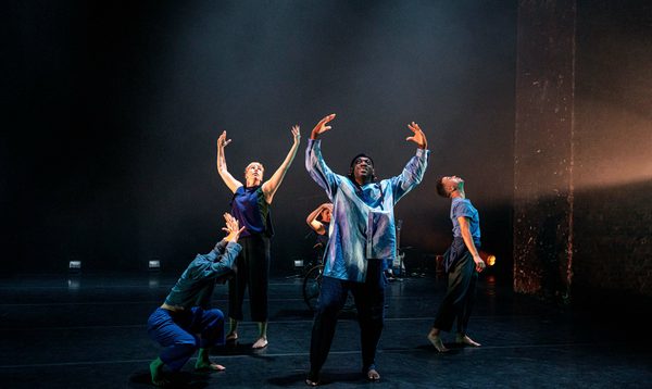 Five dancers in formation on stage, two with their arms curved above their head