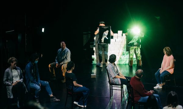 Two people stand on stage with mirrors on their back, with a group of people around them on chairs