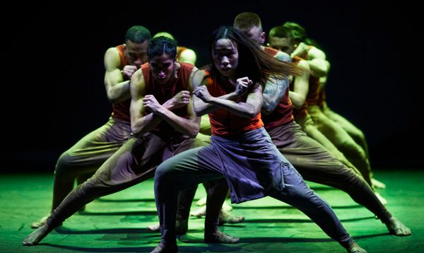 A group of dancers stand in a triangle formation with their arms in a cross in front of them