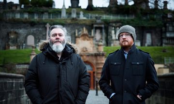 Two men stand in black coats with their hands in their pockets in front of an old stone building