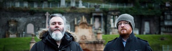 Two men stand in black coats with their hands in their pockets in front of an old stone building