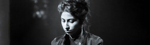 A greyscale image of a spotlit woman looking down, wearing a black jacket with her hair up.