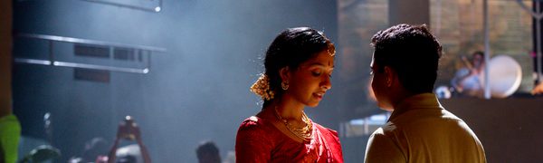 A woman in a sari and a man in a polo neck shirt stand holding hands in the foreground, with other people out of focus behind them