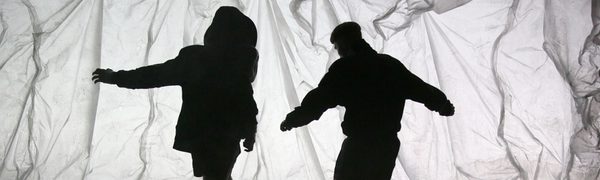 Three boys in silhouette against a translucent, crumpled background, with one of them lying on the floor and two of them standing over him.