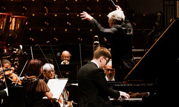 A conductor stands with his arms raised in front of him in front of an orchestra