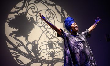 A smiling woman stands with her arms up in the air, wearing a bright blue head wrap.
