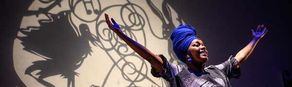 A smiling woman stands with her arms up in the air, wearing a bright blue head wrap.