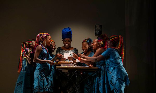 A group of women dressed in vibrant blue dresses stand around a table smiling at pieces of paper