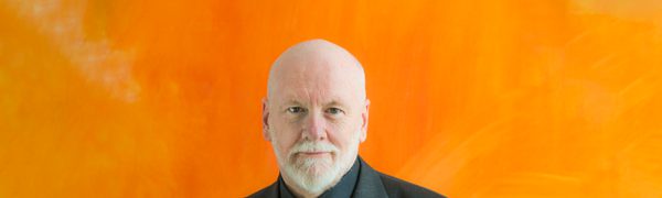 A bearded man in a black suit looks straight at the camera, sitting in front of a bright orange painting