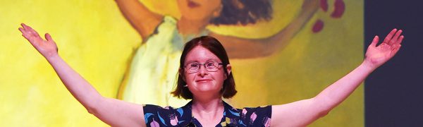A woman with Down Syndrome stands with her arms open wide in front of a painting of a child with her arms in the same position.