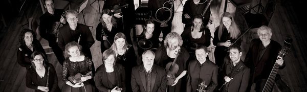 A black and white group photo of the orchestra ensemble all dressed up for concert, standing and looking up at the camera while holding their instruments in their hands